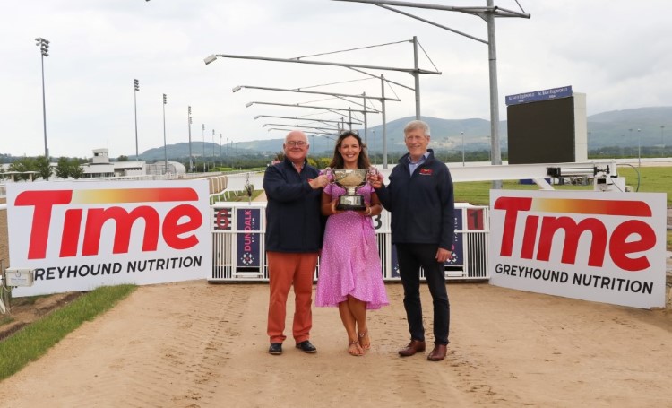 Trophy for Time Greyhound Nutrition Dundalk International This is an image of three people. A man on the left, a woman in the middle and a man on the right. All three are standing up and holding the trophy for the 2026 Dundalk International. They are standing in front of the traps on the track of Dundalk Stadium. It is a sunny day and the mountains are in the background.