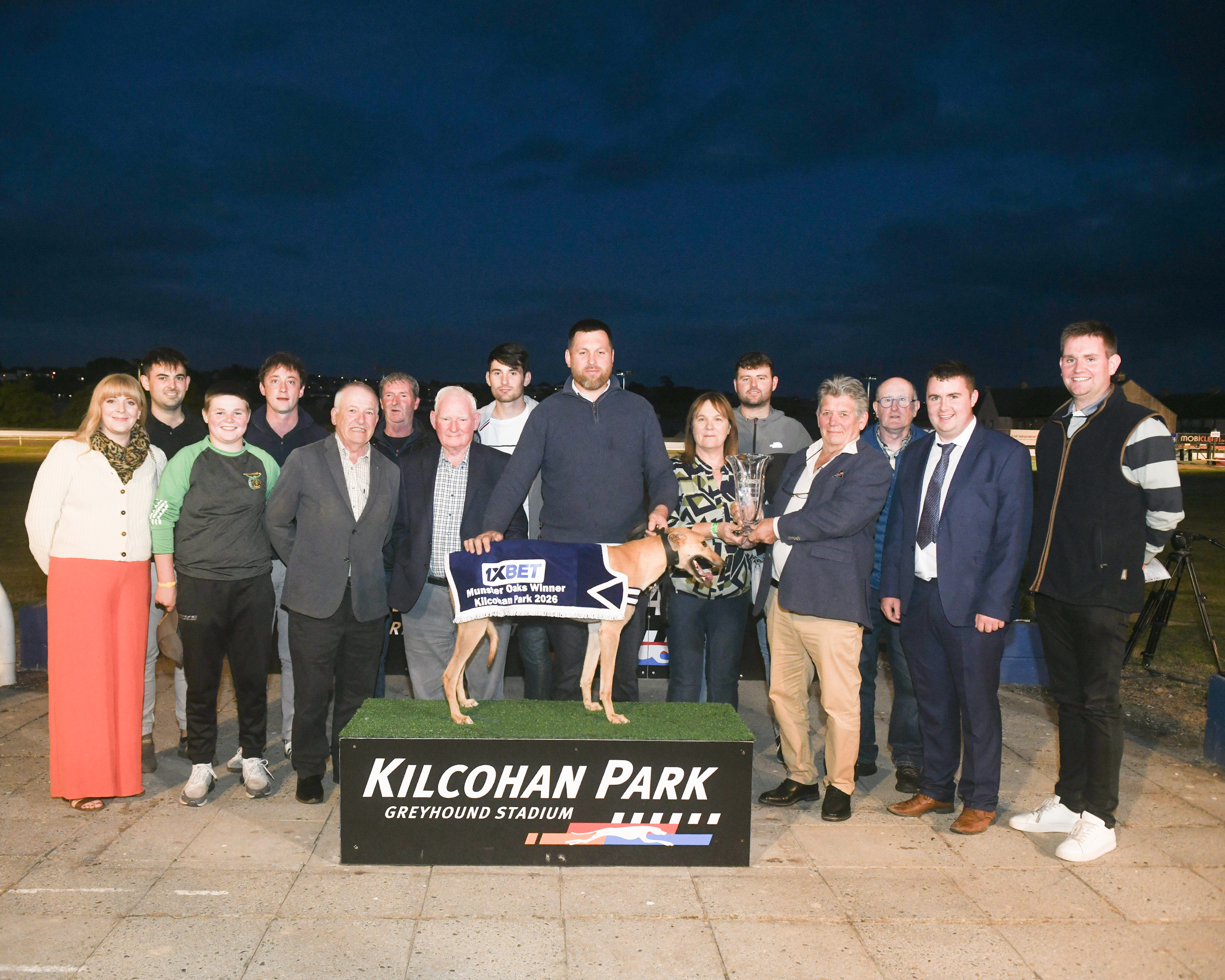 Trophy Presentation for 1XBET Munster Oaks Winner Long Siesta Image show a group presentation photograph at Kilcohan Park Greyhound Stadium showing the 1XBET Munster Oaks winner, Long Siesta, standing on a podium wearing a winner's jacket, surrounded by connections and race officials, with a trophy presentation taking place.