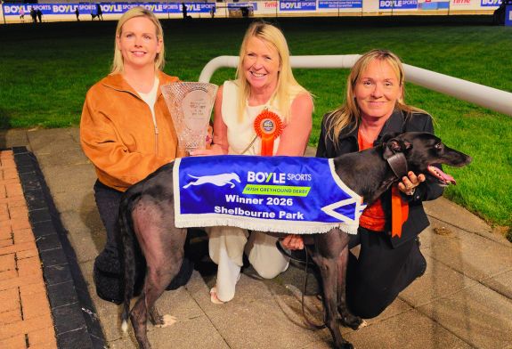 Image shows 3 women, Jenna Boyle, Bev Lochead and Nicky Holland, in the infield in Shelbourne Park kneeling behind a black greyhound, Bockos Gold, wearing the 2026 BOYLE Sports Irish Greyhound Derby winner's jacket. Jenna is handing a large pece of crystal, the winner's trophy, to Bev. 