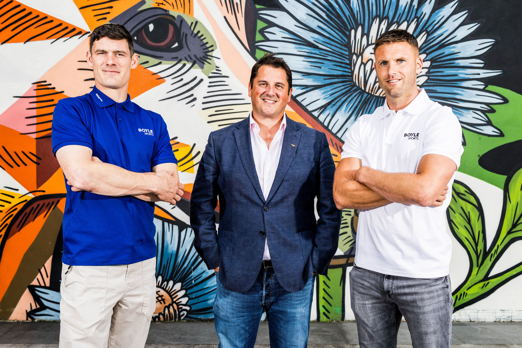 CONNOLLY VS CADOGAN: DUBLIN TAKES ON CORK IN BOYLE Sports Irish Greyhound Derby Commentator Challenge The image shows 3 men, Diarmuid Connolly, Ian Fortune and Eoin Cadogan, against a colourful mural of a greyhound outside Dublin's Shelbourne Park Greyhound Stadium to launch their Celebrity Commentator Challenge