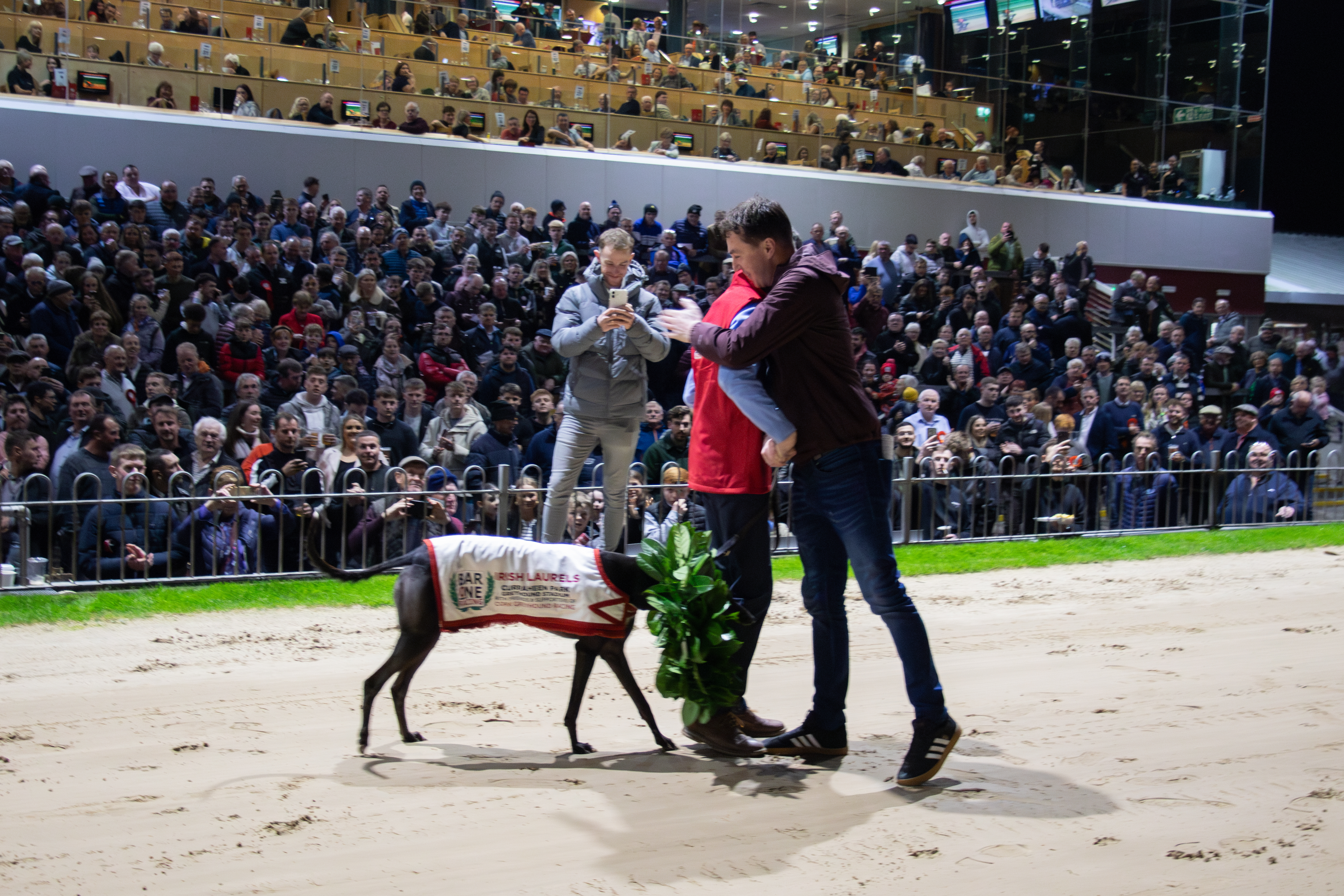 Trainer Patrick Guilfoyle hugs handler Jayden Loughnane with Magical On Fire Image shows a moment just after the 2025 Bar One Racing Irish Laurels Final where trainer Patrick Guilfoyle embraces Jayden Loughnane in celebration as he leads greyhound Magical Mag along the sand track back to the winner's podium in front of a very packed stand in Cork's Curraheen Park Greyhound Stadium